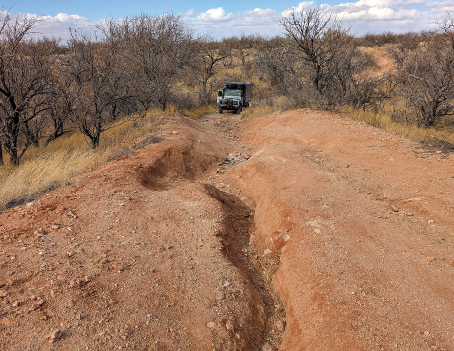 Off-road vehicle approaching a deeply eroded dirt trail in a dry, rugged landscape with sparse leafless trees and patches of grass under a partly cloudy sky