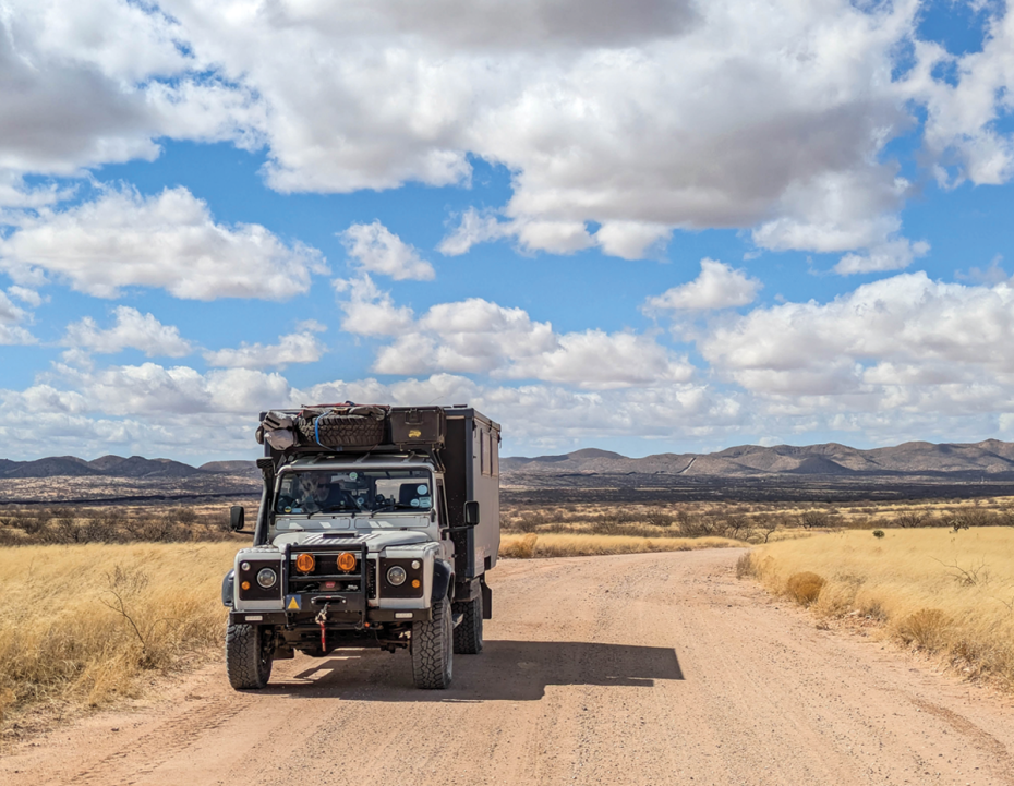 Expedition vehicle driving along a wide dirt road in an open desert landscape, surrounded by dry grass and distant hills under a bright blue sky with scattered white clouds