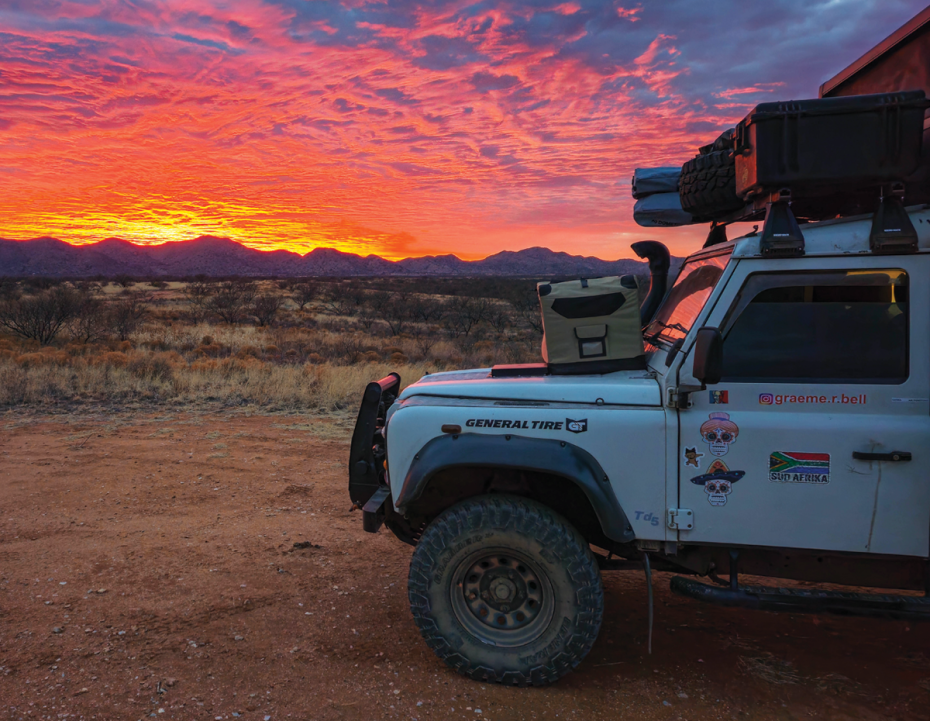 Off-road expedition vehicle parked on desert terrain at sunset, with a vivid orange and pink sky illuminating distant mountains and dry grassland.